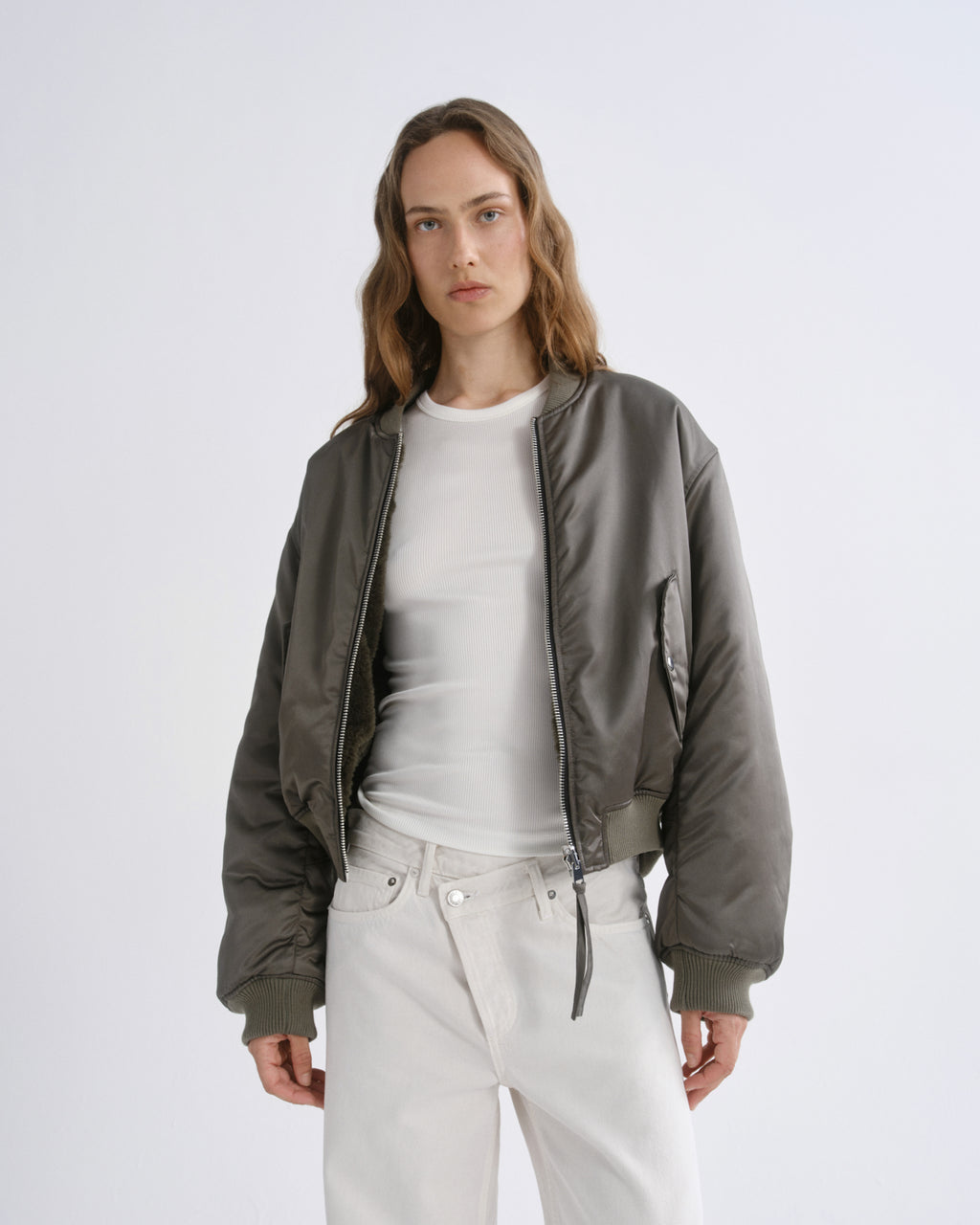 A woman modeling a olive green bomber jacket over a white t-shirt and white pants, photographed against a grey background.