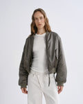 A woman modeling a olive green bomber jacket over a white t-shirt and white pants, photographed against a grey background.