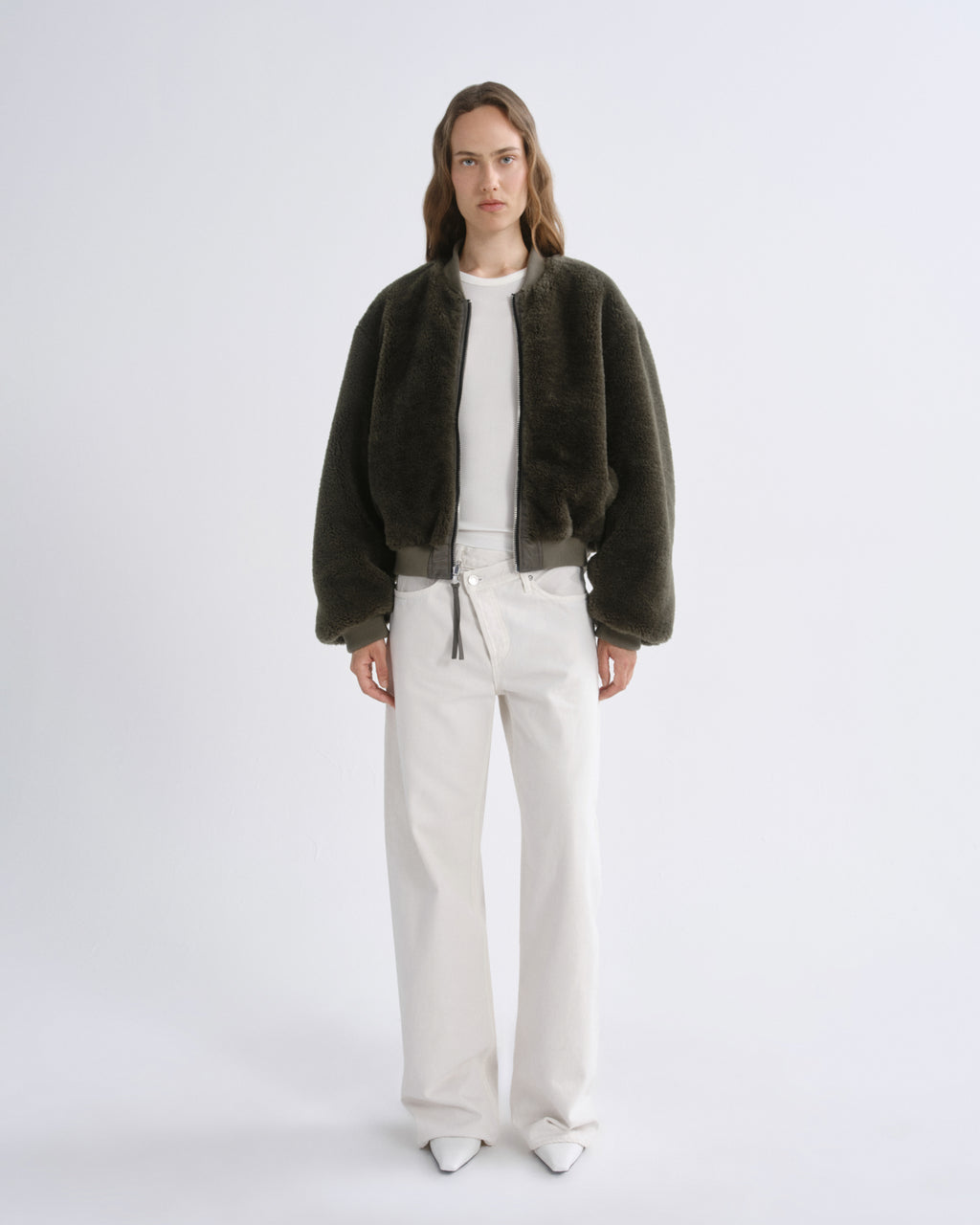 A model stands against a grey background wearing a dark olive green fuzzy cropped jacket over a white top and white wide-leg pants.
