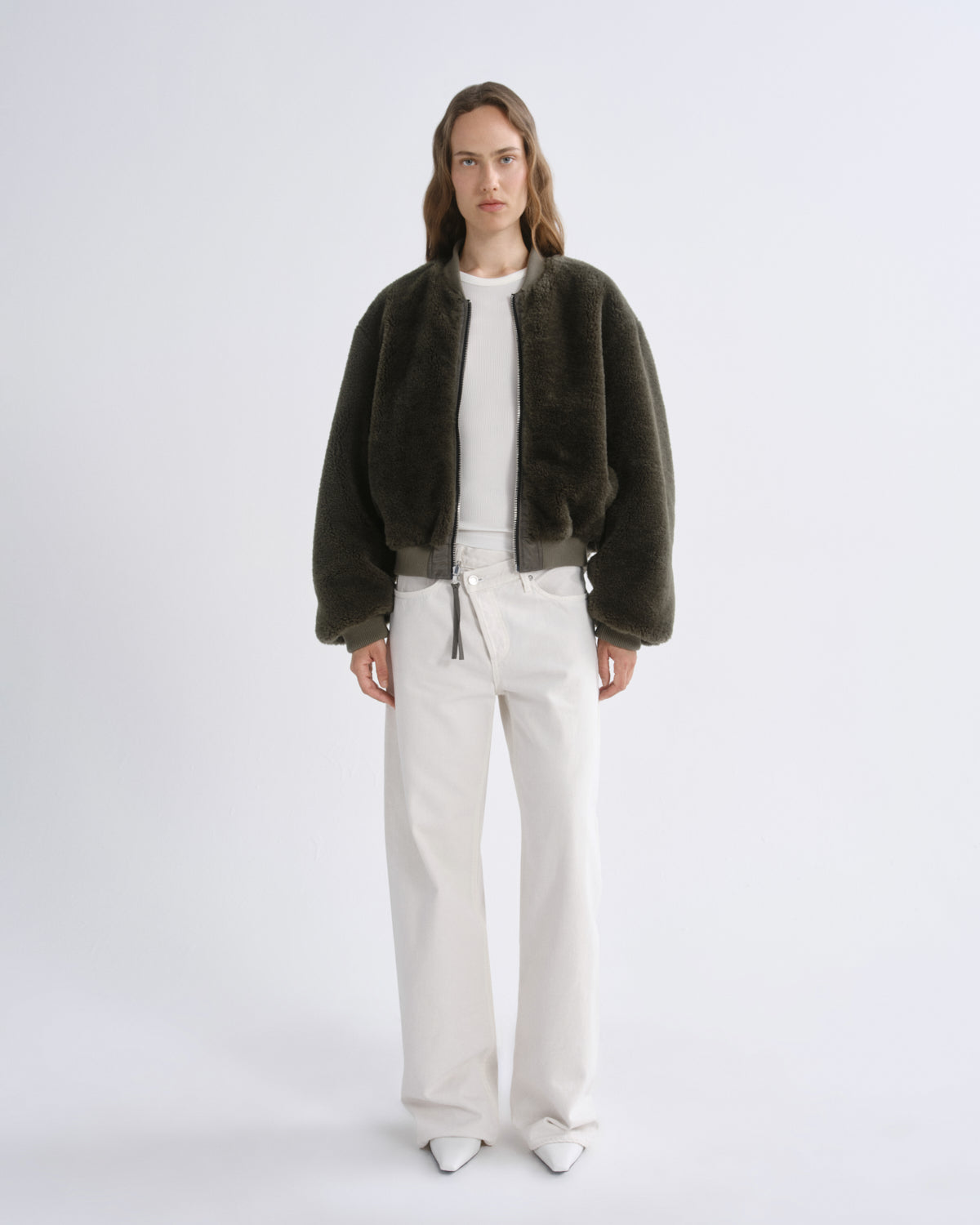 A model stands against a grey background wearing a dark olive green fuzzy cropped jacket over a white top and white wide-leg pants.