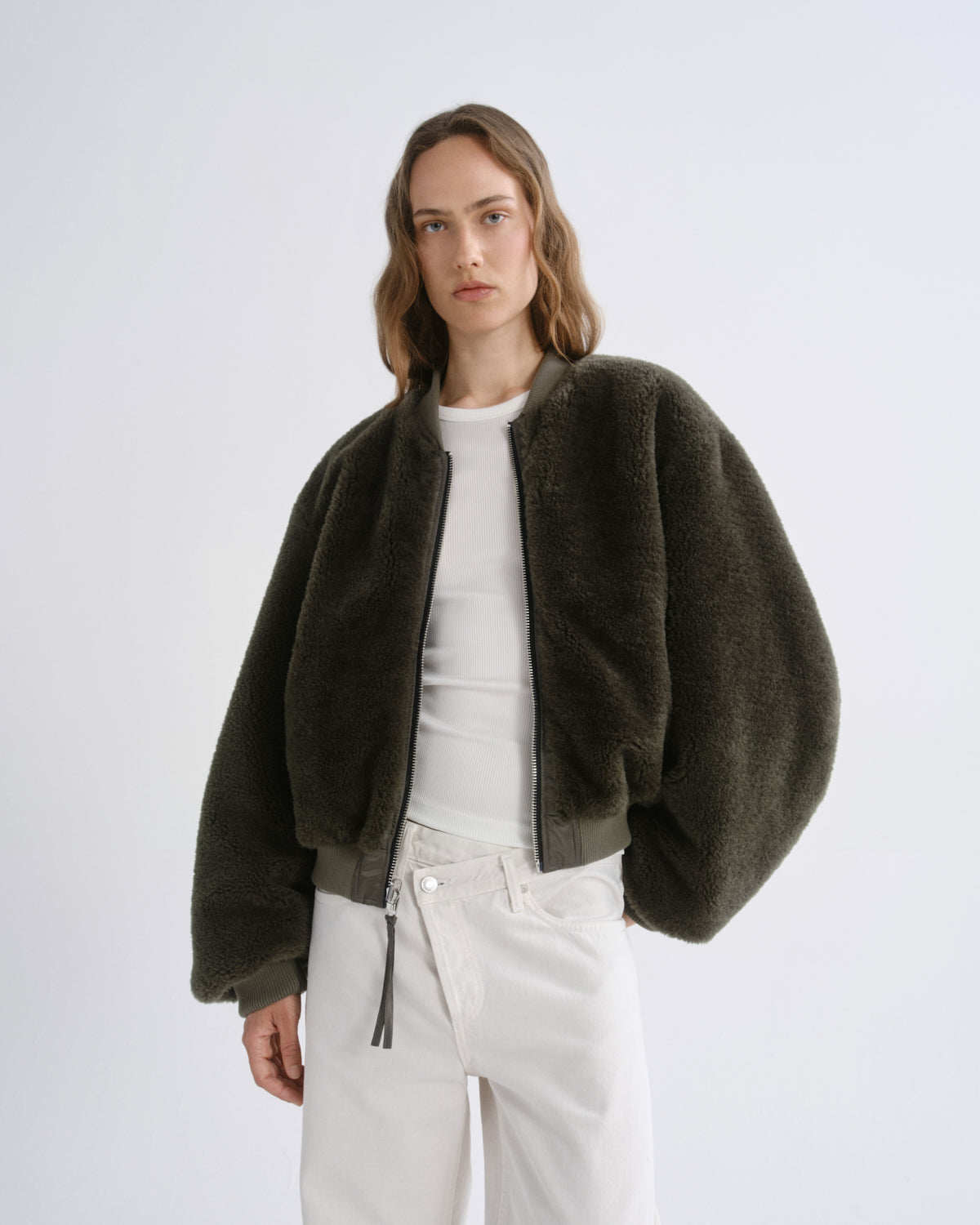 A model wearing a dark olive green fuzzy cropped jacket over a white top and white pants, photographed against a grey background.