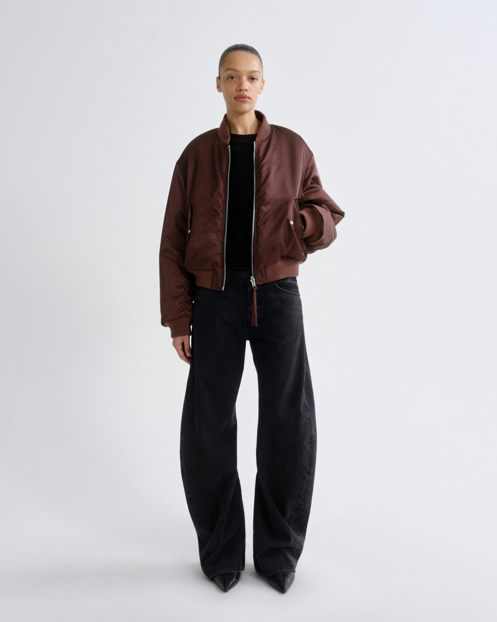 Person wearing a brown red bomber jacket and black pants on a grey background