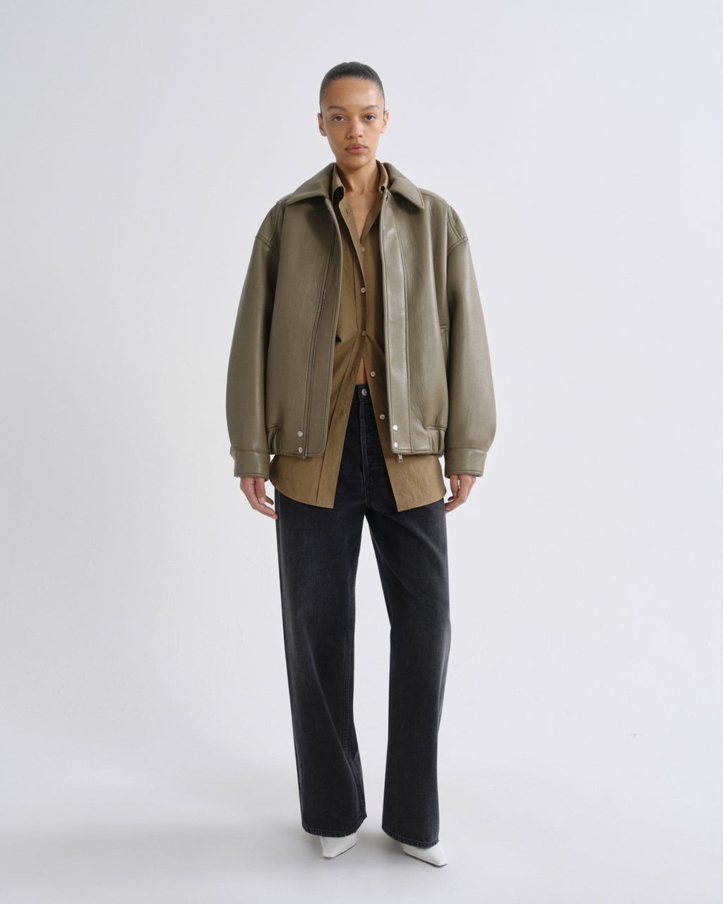 A model stands against a light gray studio background wearing an oversized olive green jacket over a tan shirt, paired with dark wide-leg jeans and white pointed shoes.