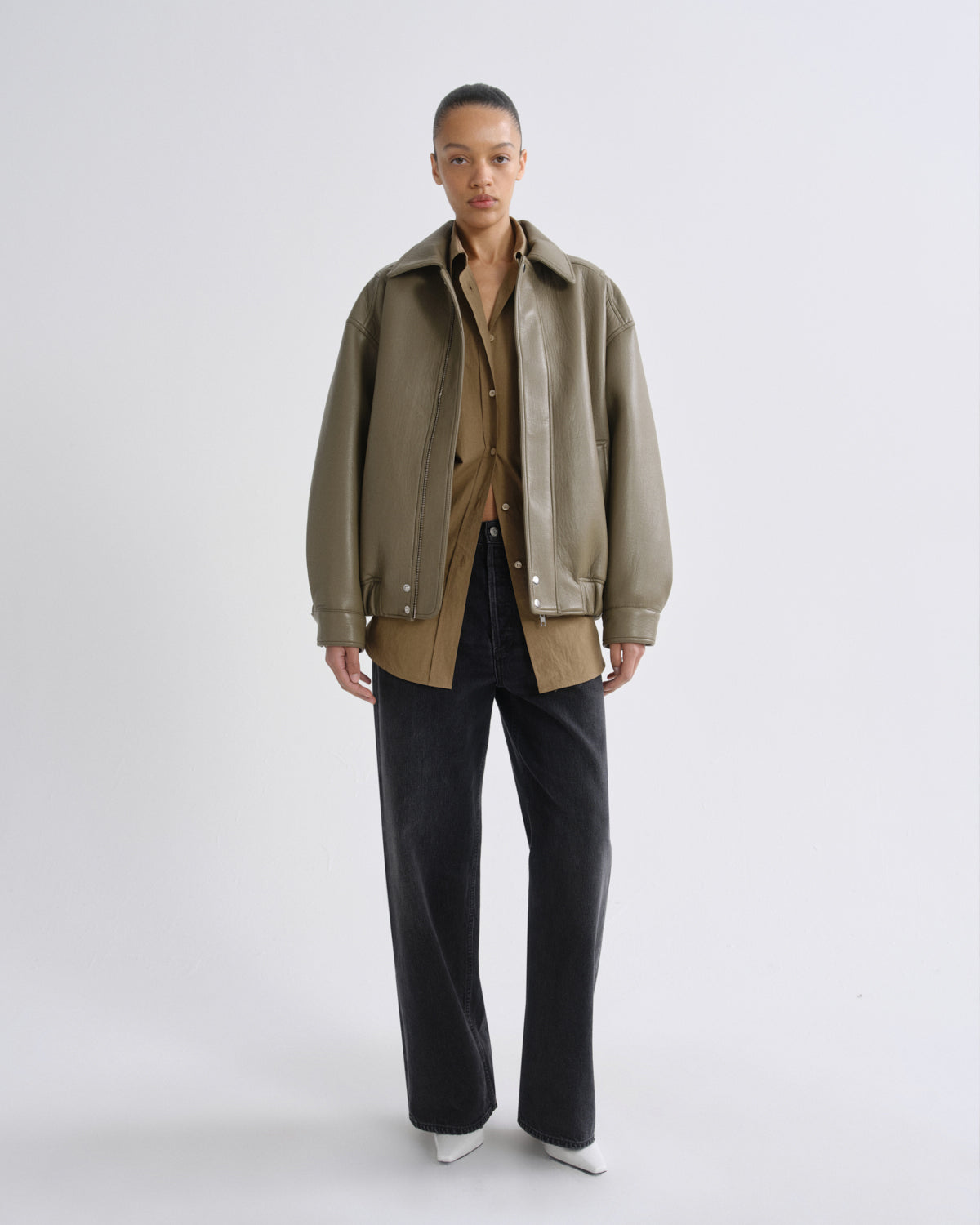 A model stands against a light gray studio background wearing an oversized olive green jacket over a tan shirt, paired with dark wide-leg jeans and white pointed shoes.