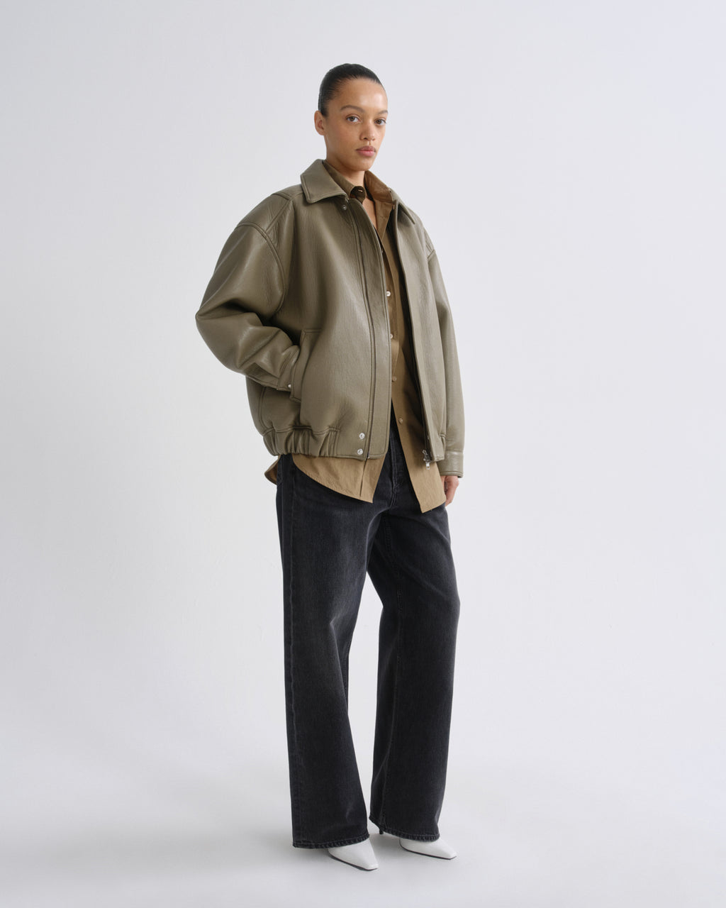A model stands against a light gray studio background wearing an oversized olive green leather jacket over a tan shirt, paired with dark wide-leg jeans and white pointed shoes.