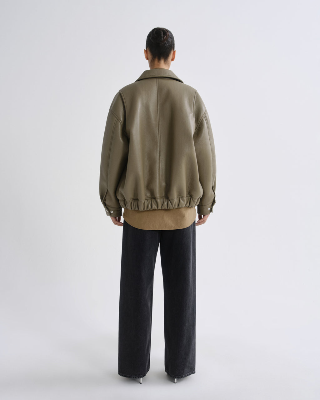 A model viewed from the back wearing an oversized olive green leather jacket over a tan shirt and black wide-leg trousers, shot against a light gray studio background.