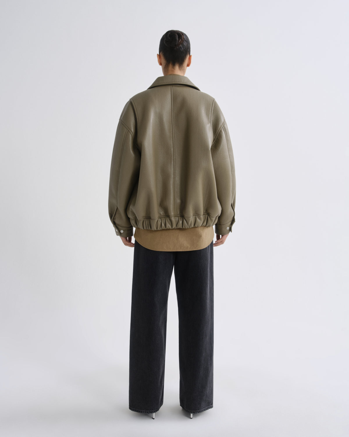 A model viewed from the back wearing an oversized olive green leather jacket over a tan shirt and black wide-leg trousers, shot against a light gray studio background.