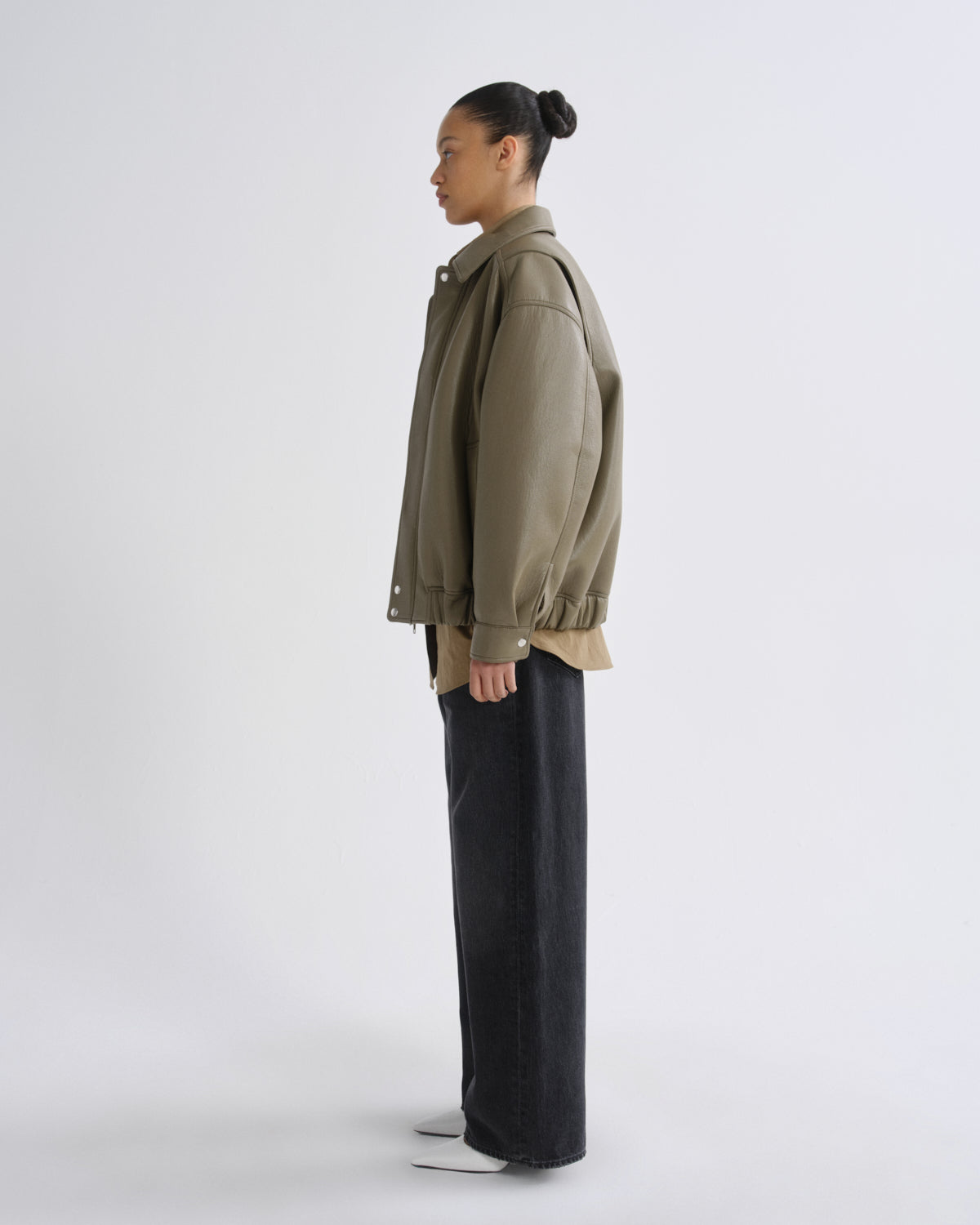 A model stands in profile wearing an oversized olive green utility jacket over a tan shirt, paired with dark charcoal wide-leg pants and white shoes, against a plain light gray studio background.