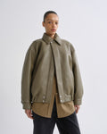 A model wearing an oversized olive green faux-leather jacket with a collar and snap-button front, layered over a tan shirt and dark pants, against a plain light gray studio background.