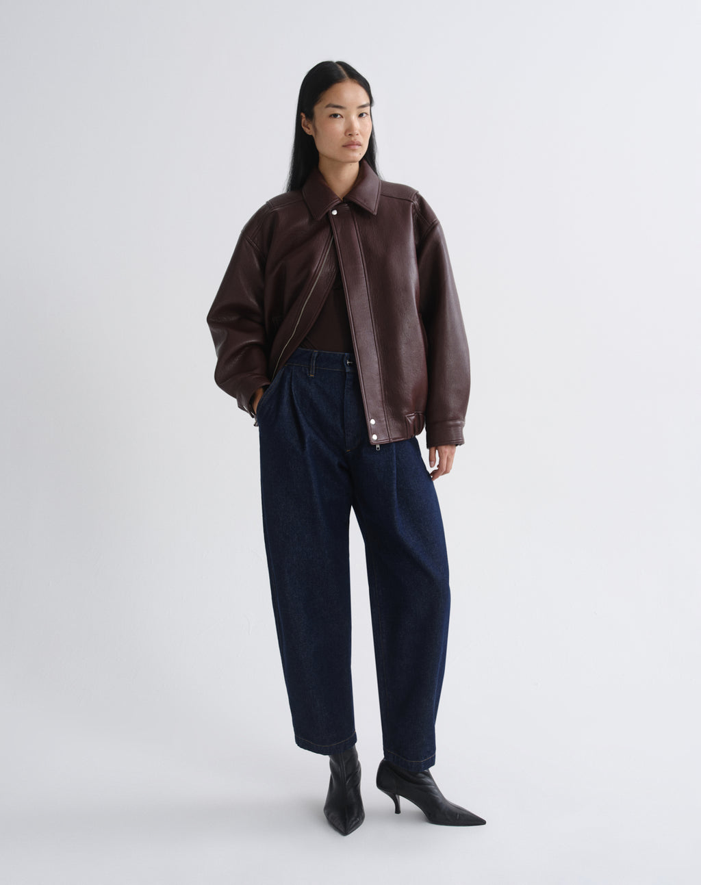 A model stands against a grey background wearing a dark brown leather jacket, dark blue wide-leg jeans, and black pointed ankle boots.
