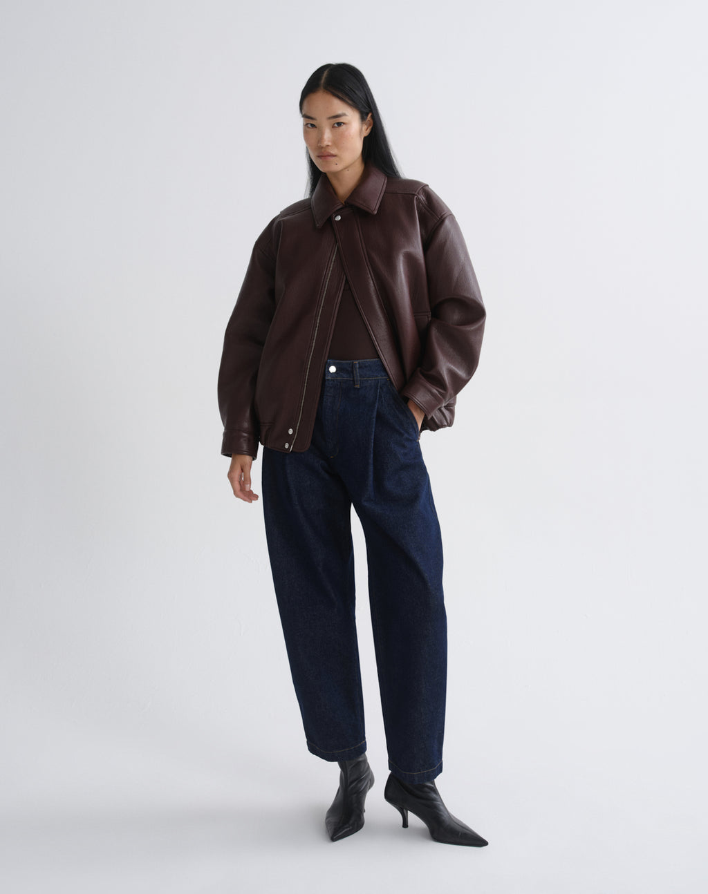A model stands against a grey background wearing a dark brown leather jacket, dark blue wide-leg jeans, and black pointed-toe heeled boots.