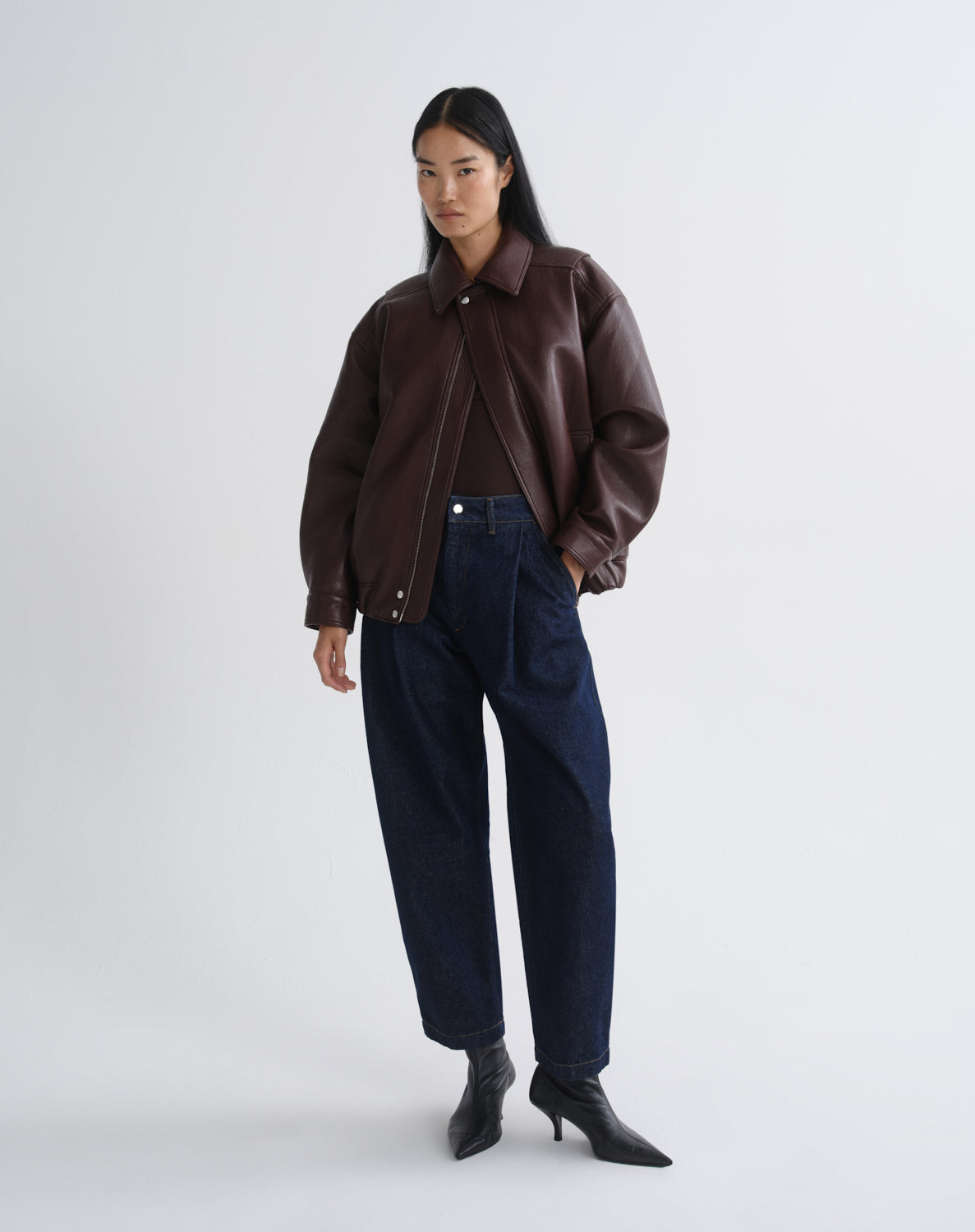 A model stands against a grey background wearing a dark brown leather jacket, dark blue wide-leg jeans, and black pointed-toe heeled boots.
