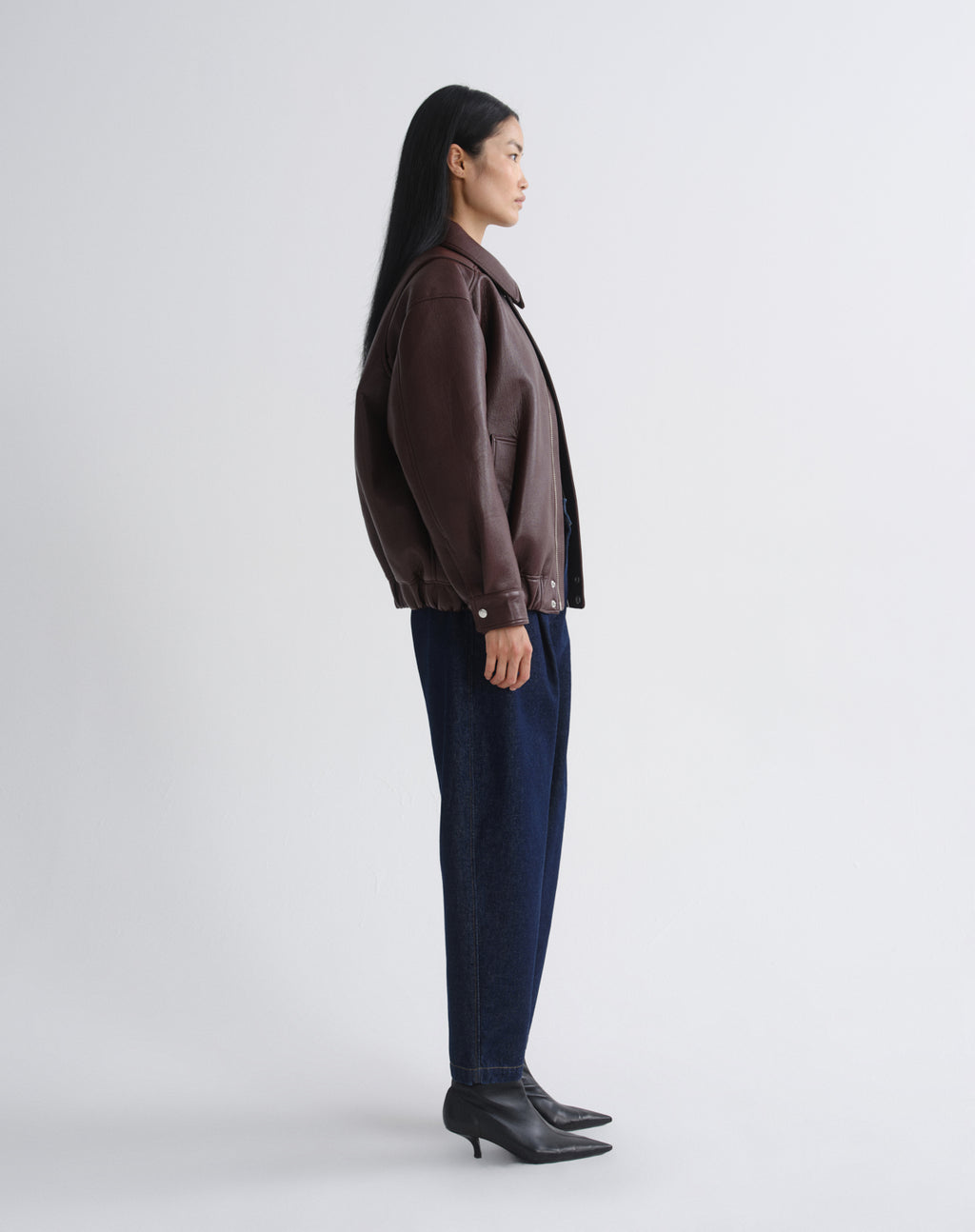 A woman shown in profile wearing a dark brown leather jacket, dark blue jeans, and black ankle boots against a grey background.