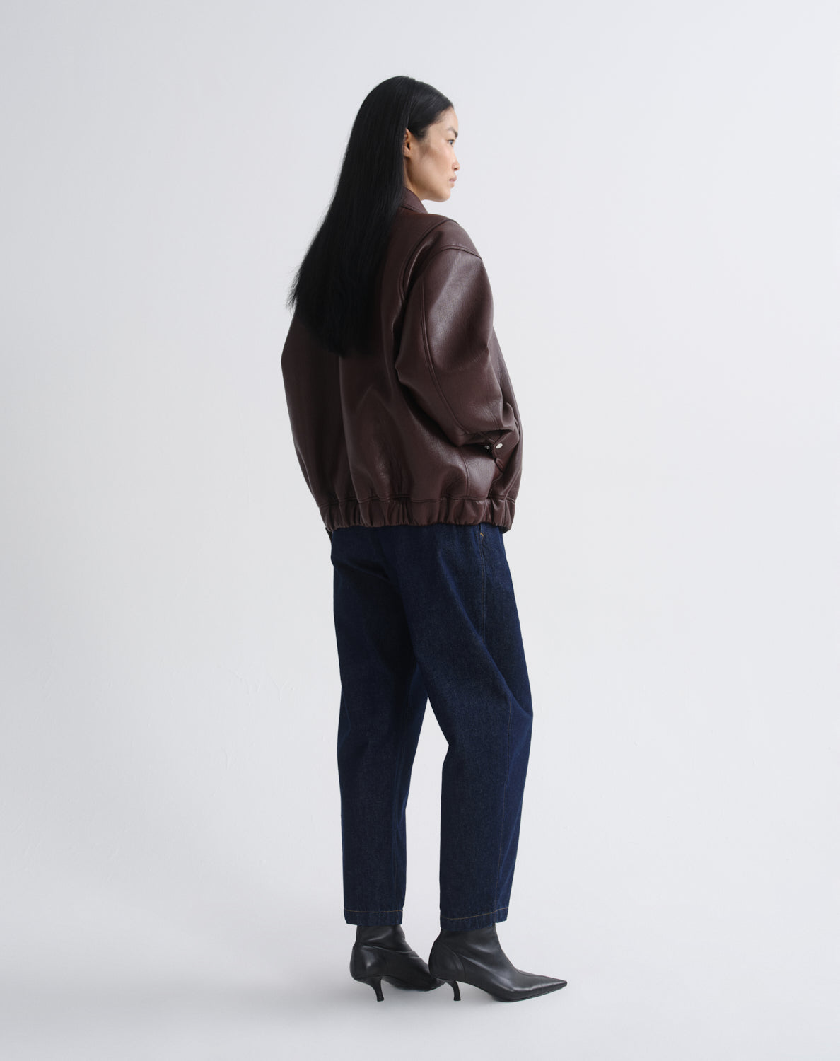 A woman shown from the back wearing a dark brown leather jacket, dark blue jeans, and black pointed-toe ankle boots, standing against at grey background.