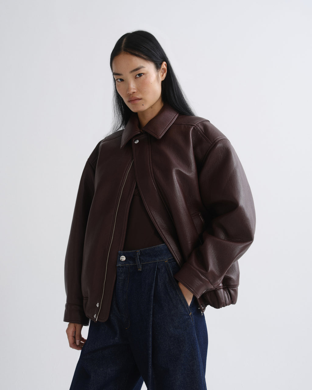 A model wearing a dark brown leather jacket with a collar and button closure, styled over a dark top and dark blue jeans against a grey background.
