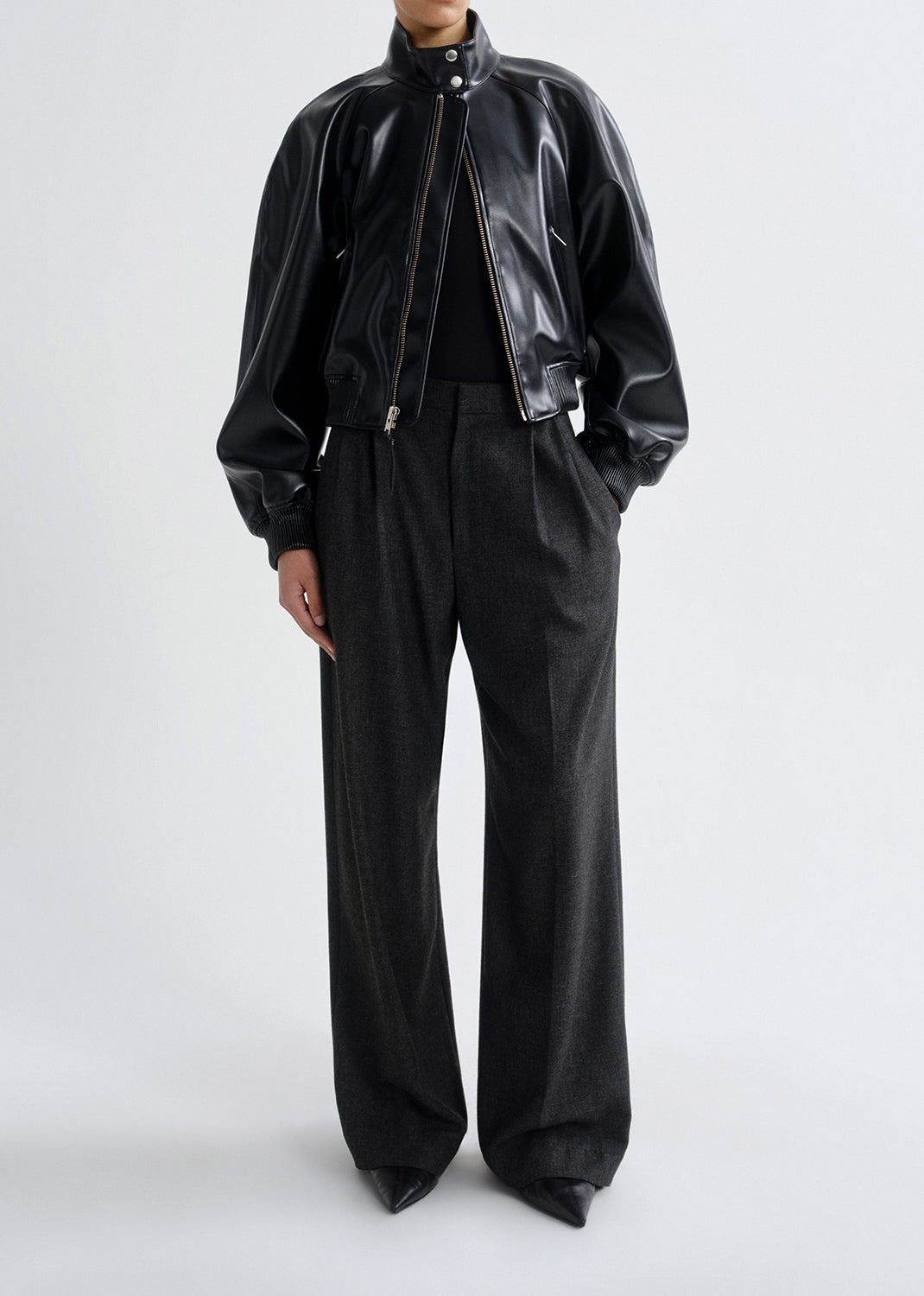 Person wearing a black leather bomber jacket with charcoal wide leg pants on a grey background.