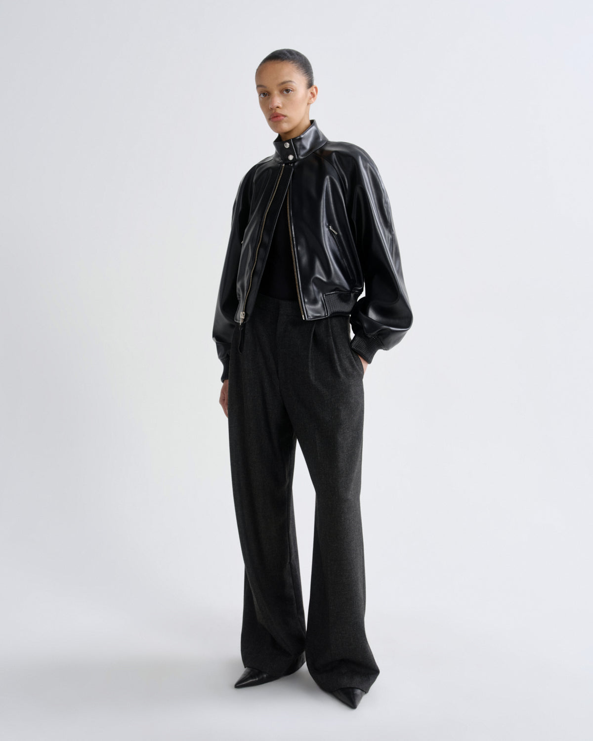 A model stands against a light gray studio background wearing a black glossy cropped jacket with a high collar, black wide-leg trousers, and black pointed shoes.