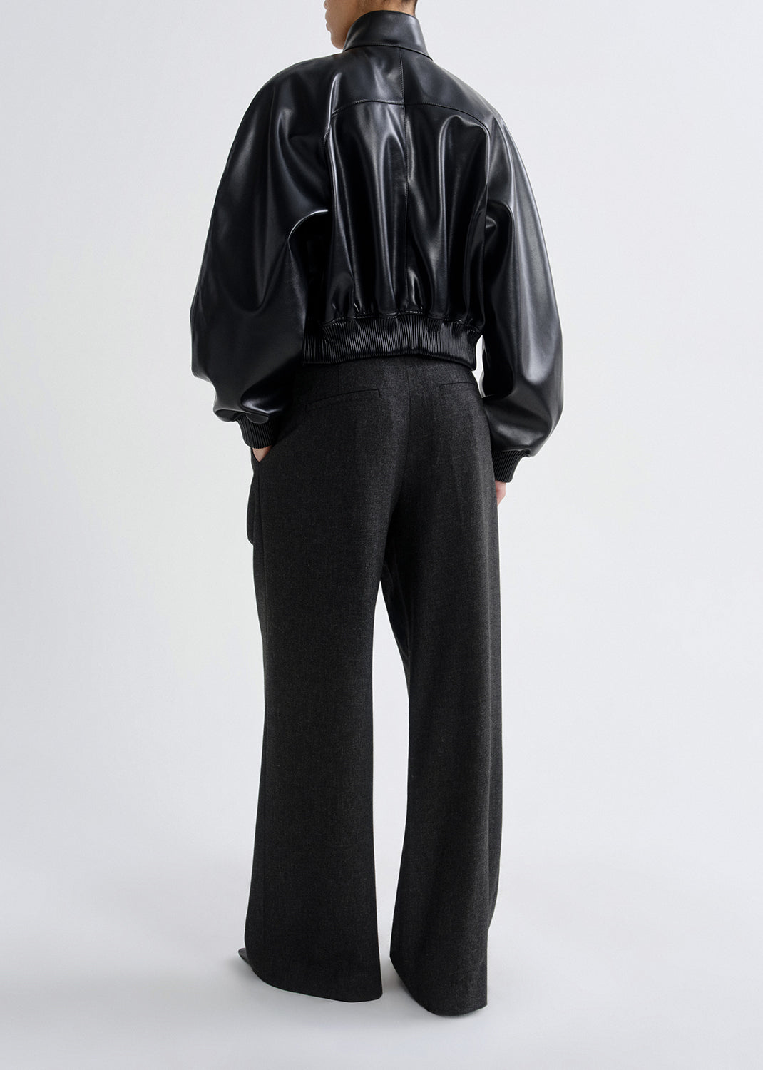 Person wearing a black leather bomber jacket with charcoal wide leg pants viewed from the back on a grey background.