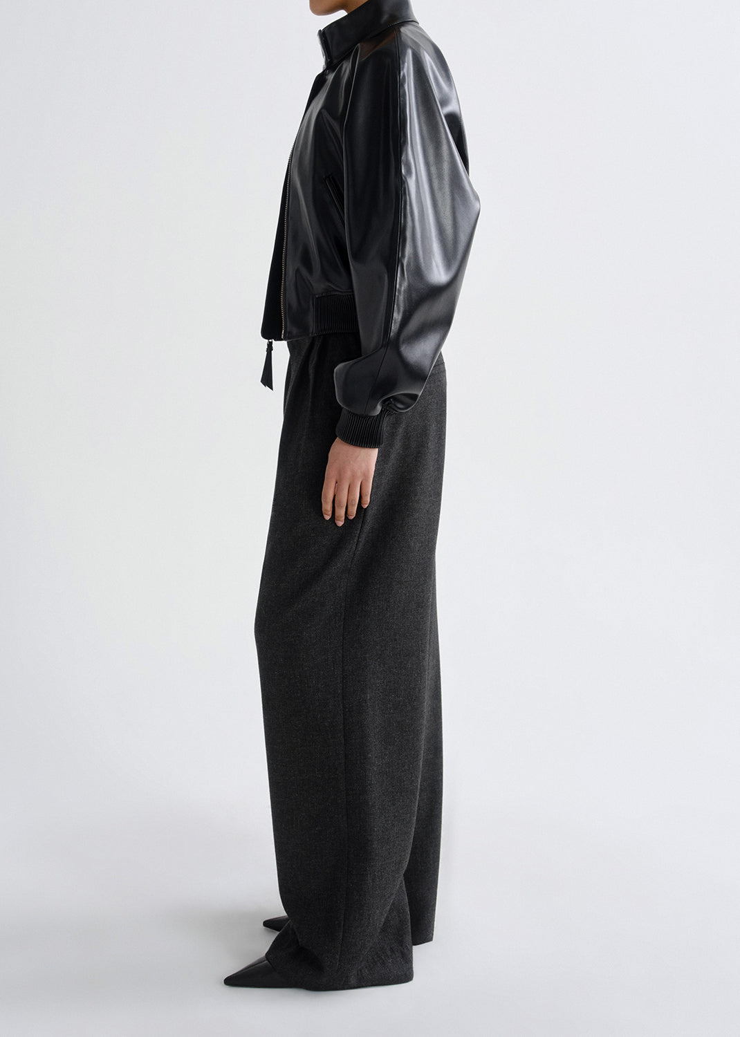 Person wearing a black leather bomber jacket with charcoal wide leg pants viewed from the side on a grey background.
