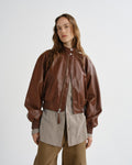 A woman modeling a brown leather bomber jacket over a layered beige and brown outfit, photographed against a clean light gray studio background.
