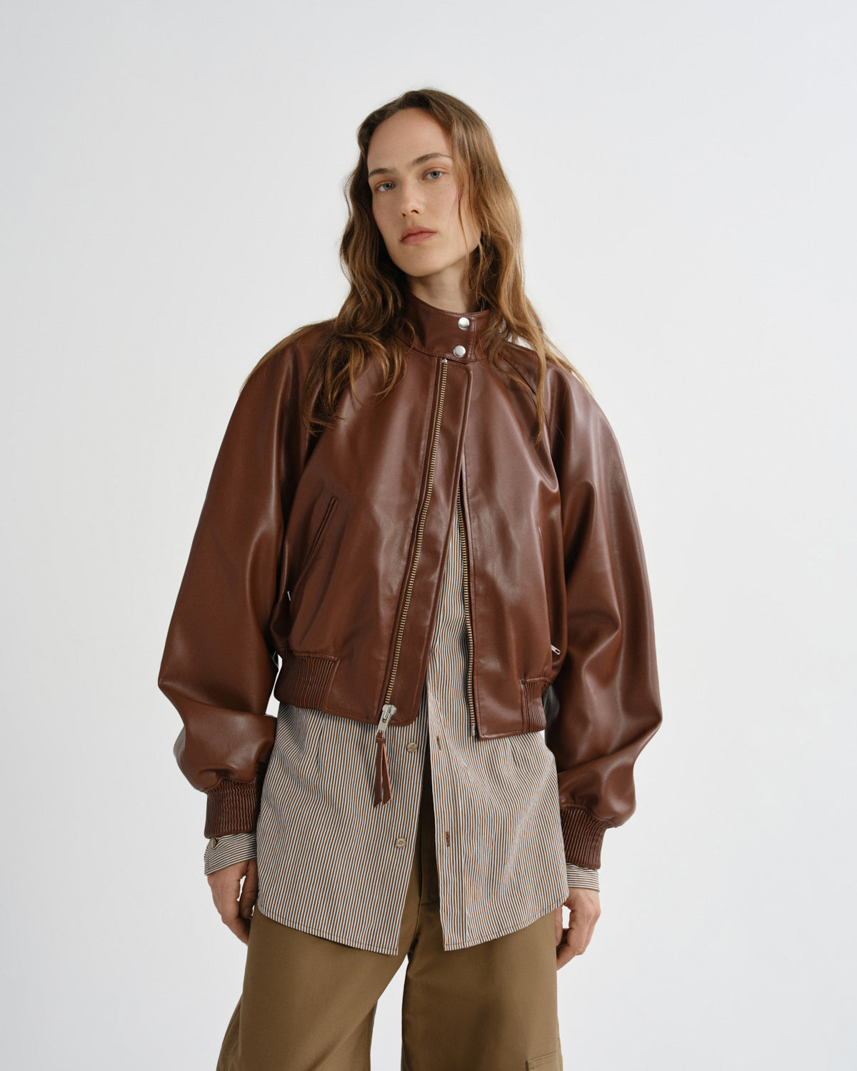 A woman modeling a brown leather bomber jacket over a layered beige and brown outfit, photographed against a clean light gray studio background.