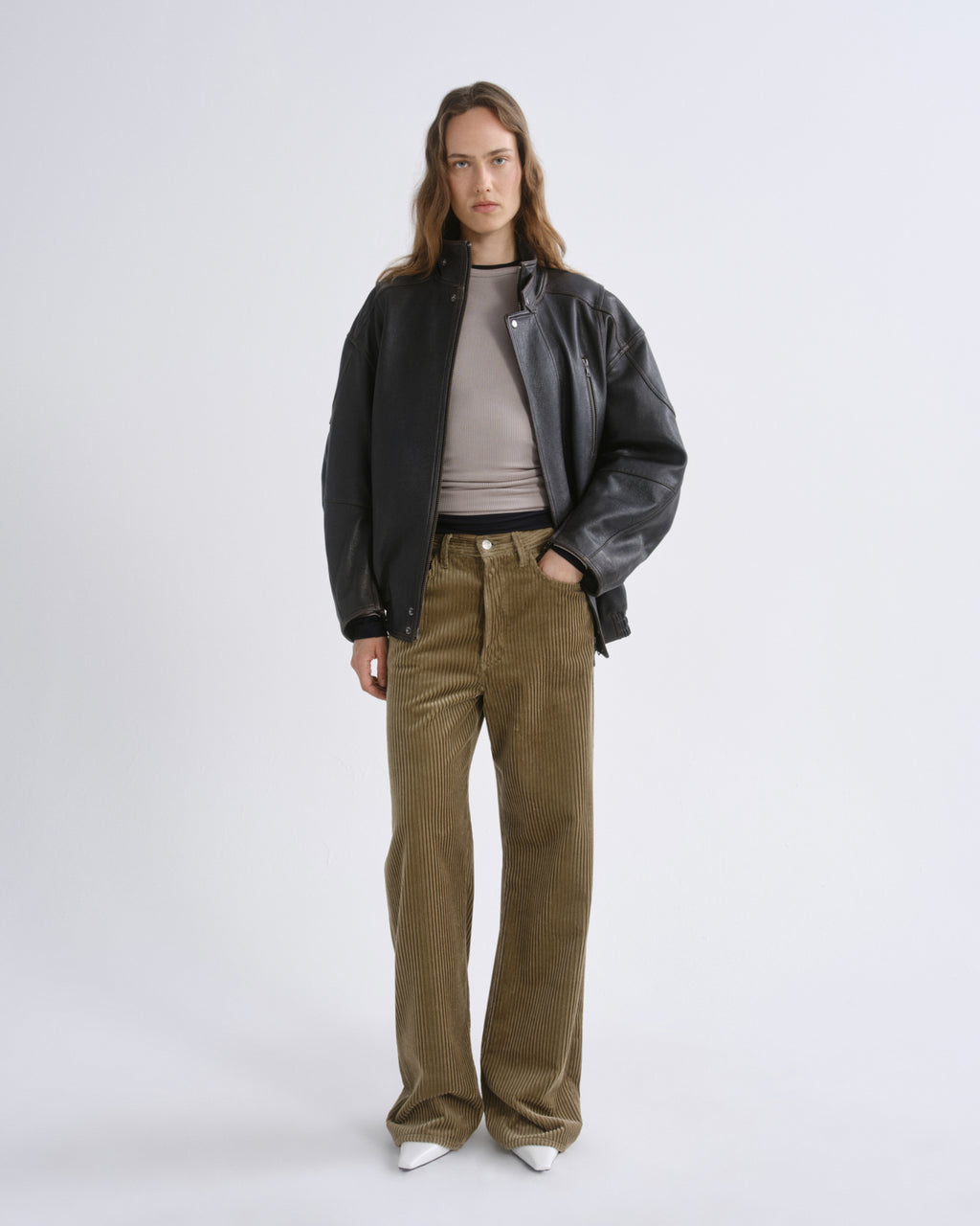 A model stands against a grey background wearing a black leather jacket over a taupe ribbed top, olive corduroy trousers, and white pointed shoes.