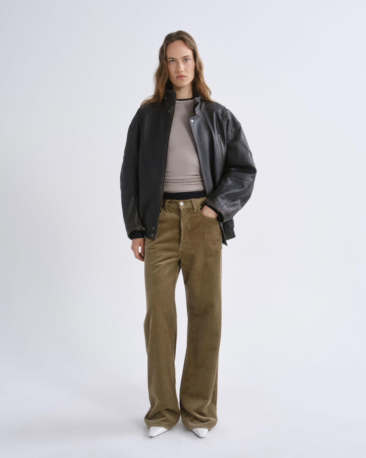 A model stands against a grey background wearing a black leather jacket over a taupe ribbed top, olive corduroy trousers, and white pointed shoes.