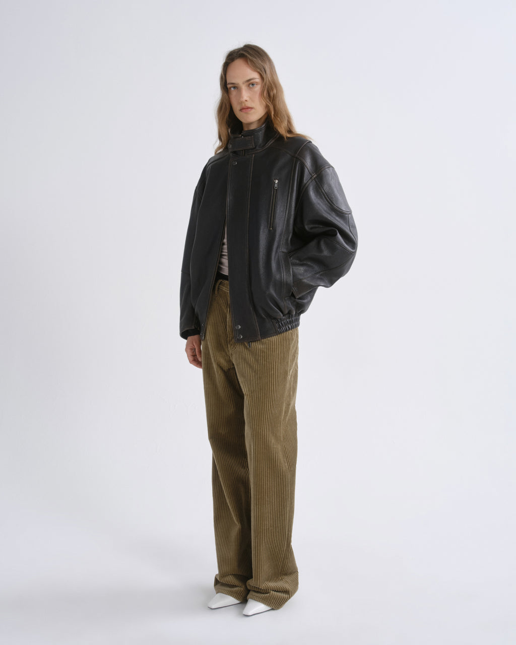 A model stands against a grey background wearing a black leather jacket, olive-brown corduroy trousers, and white pointed shoes.
