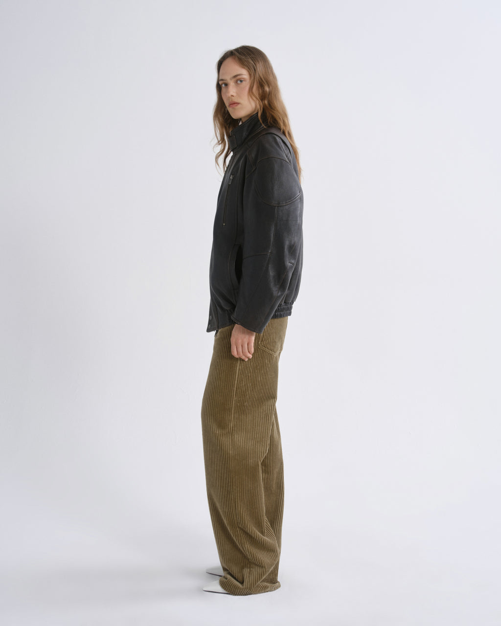 A model is shown in profile wearing a black cropped jacket and wide-leg olive corduroy trousers against a grey background.