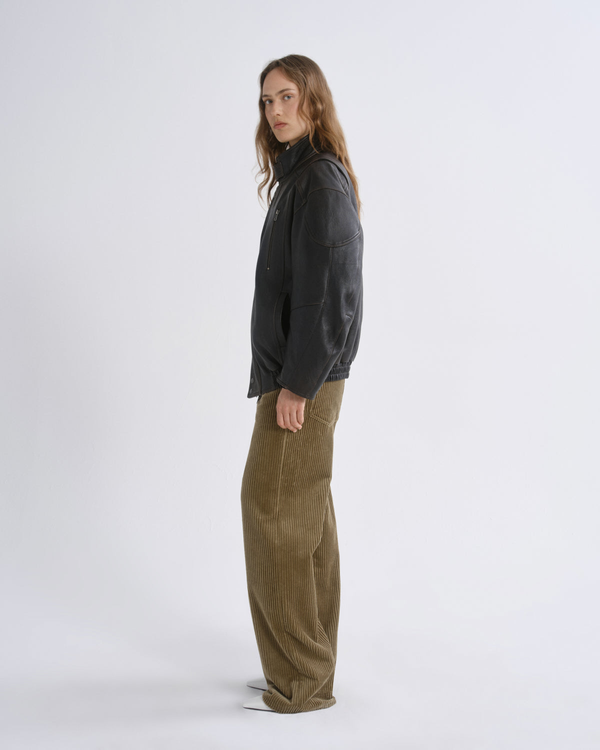 A model is shown in profile wearing a black cropped jacket and wide-leg olive corduroy trousers against a grey background.