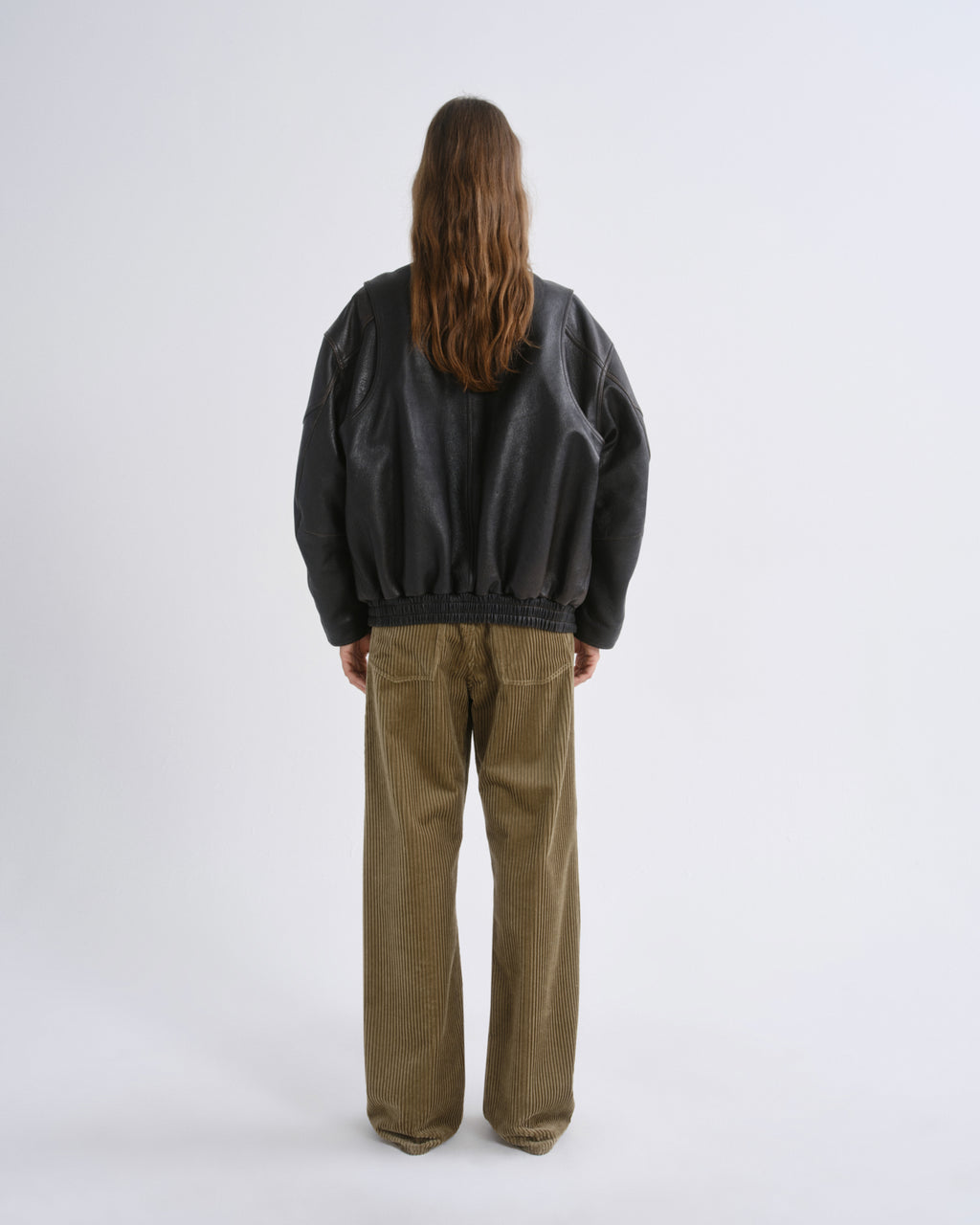A model viewed from behind wearing a black leather bomber jacket and wide-leg olive corduroy trousers, standing against a grey background.