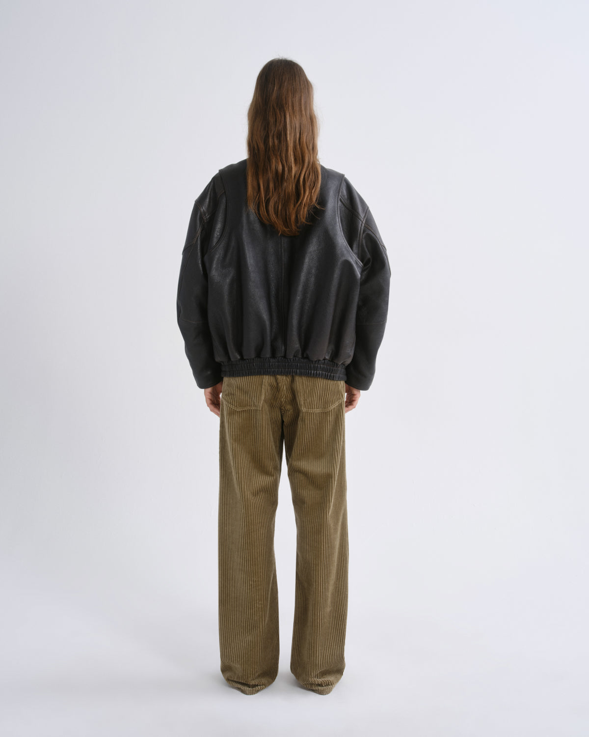A model viewed from behind wearing a black leather bomber jacket and wide-leg olive corduroy trousers, standing against a grey background.