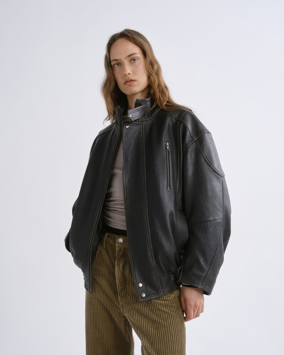 A model wearing a black leather jacket with a high collar, zippered chest pocket, and silver snap details, styled over a gray top and brown corduroy trousers, against a grey background.