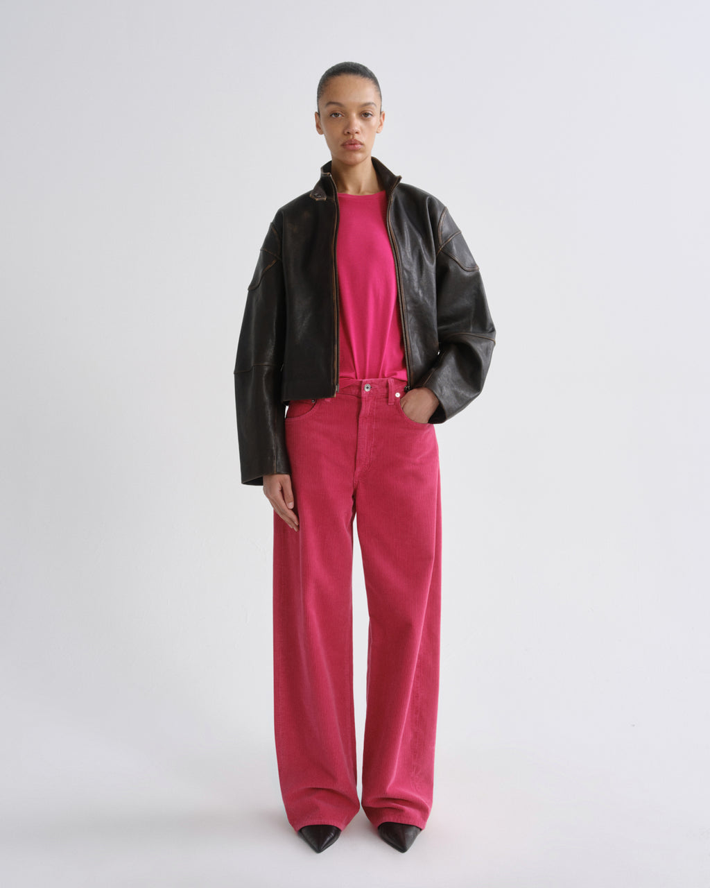 A model wearing a black leather moto jacket over a bright pink top and matching pink corduroy pants, standing against a light gray studio background.