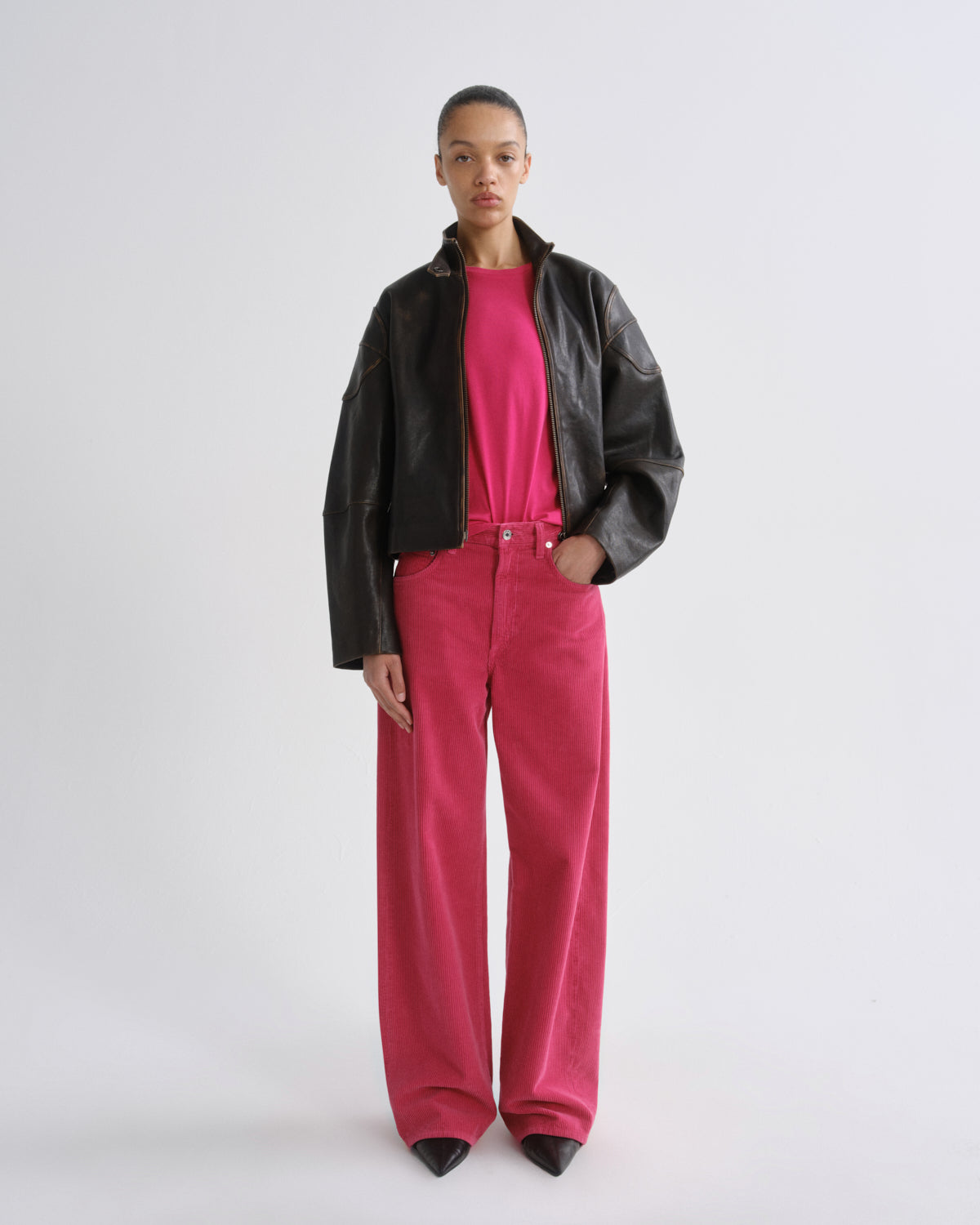 A model wearing a black leather moto jacket over a bright pink top and matching pink corduroy pants, standing against a light gray studio background.