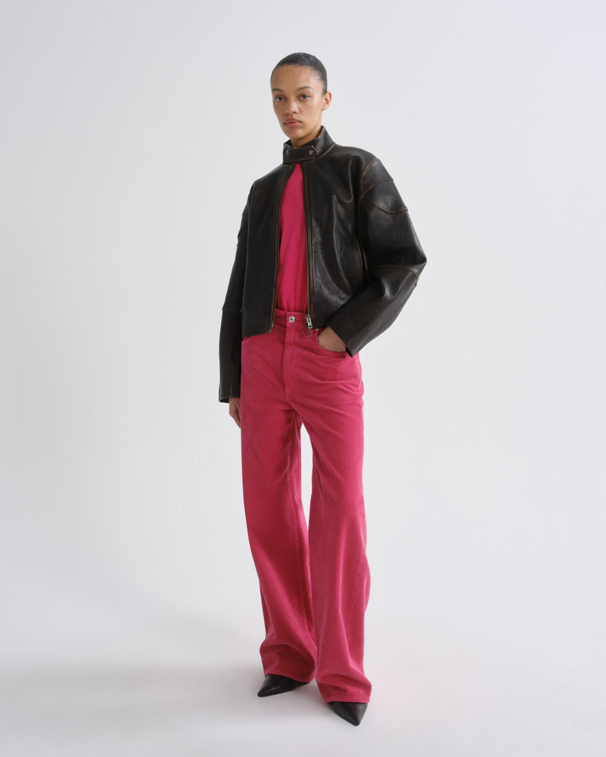A model stands against a light gray studio background wearing a black leather jacket, a bright pink shirt, and matching pink trousers.