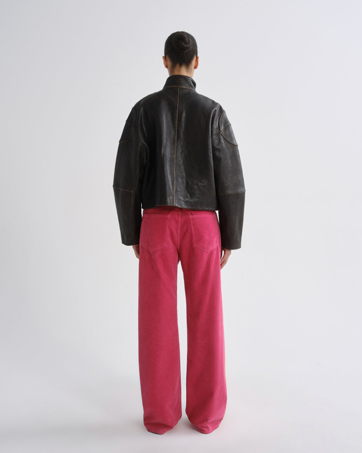 A model shown from the back wearing a black leather cropped jacket and bright pink wide-leg trousers, standing against a plain light gray studio background.