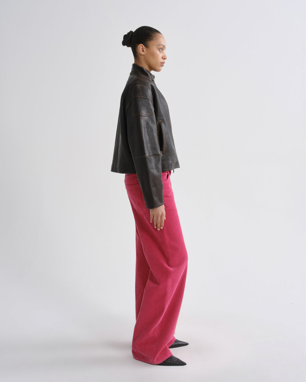 A model shown in profile wearing a dark brown leather jacket and bright pink wide-leg trousers, standing against a plain light gray studio background.