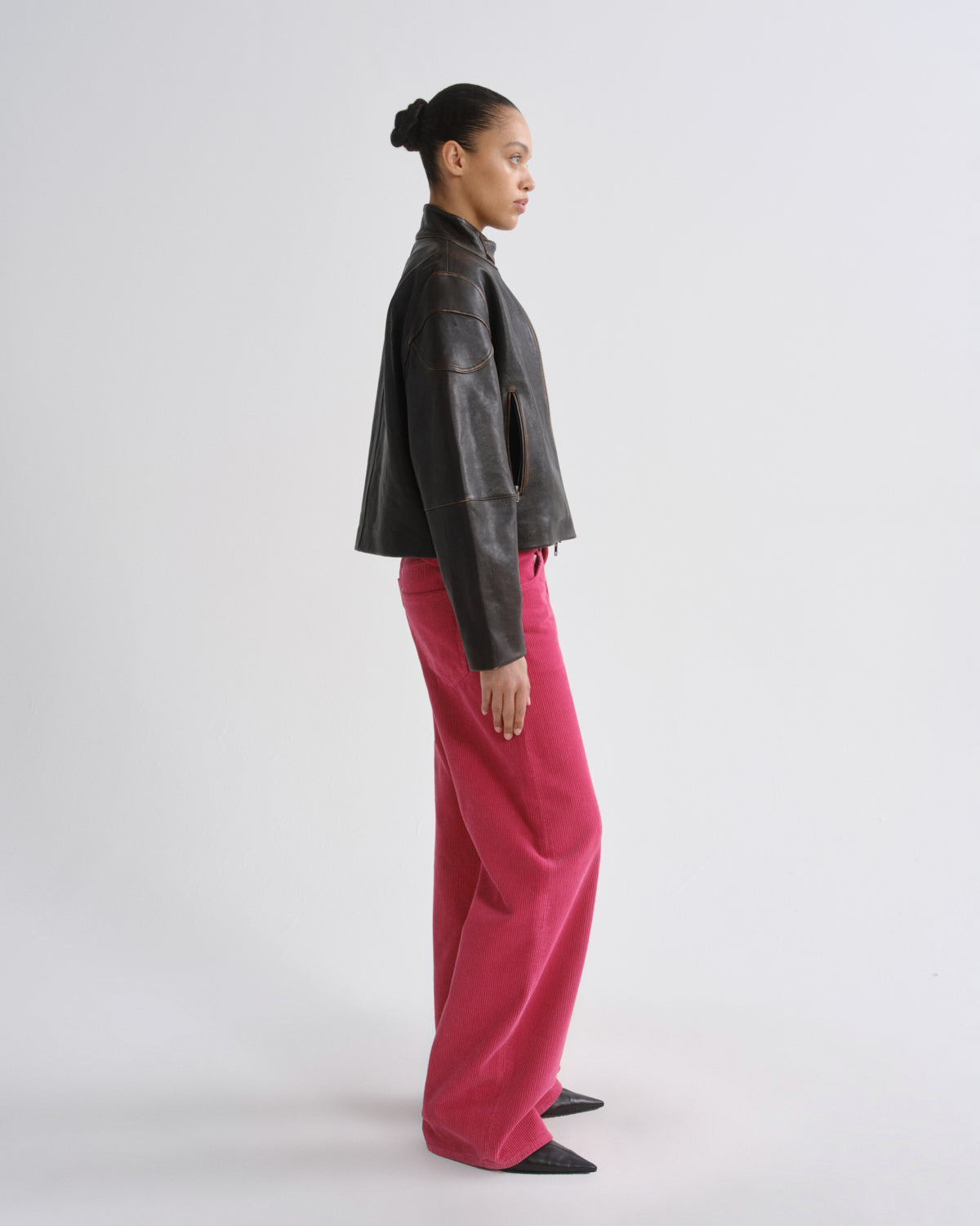 A model shown in profile wearing a dark brown leather jacket and bright pink wide-leg trousers, standing against a plain light gray studio background.