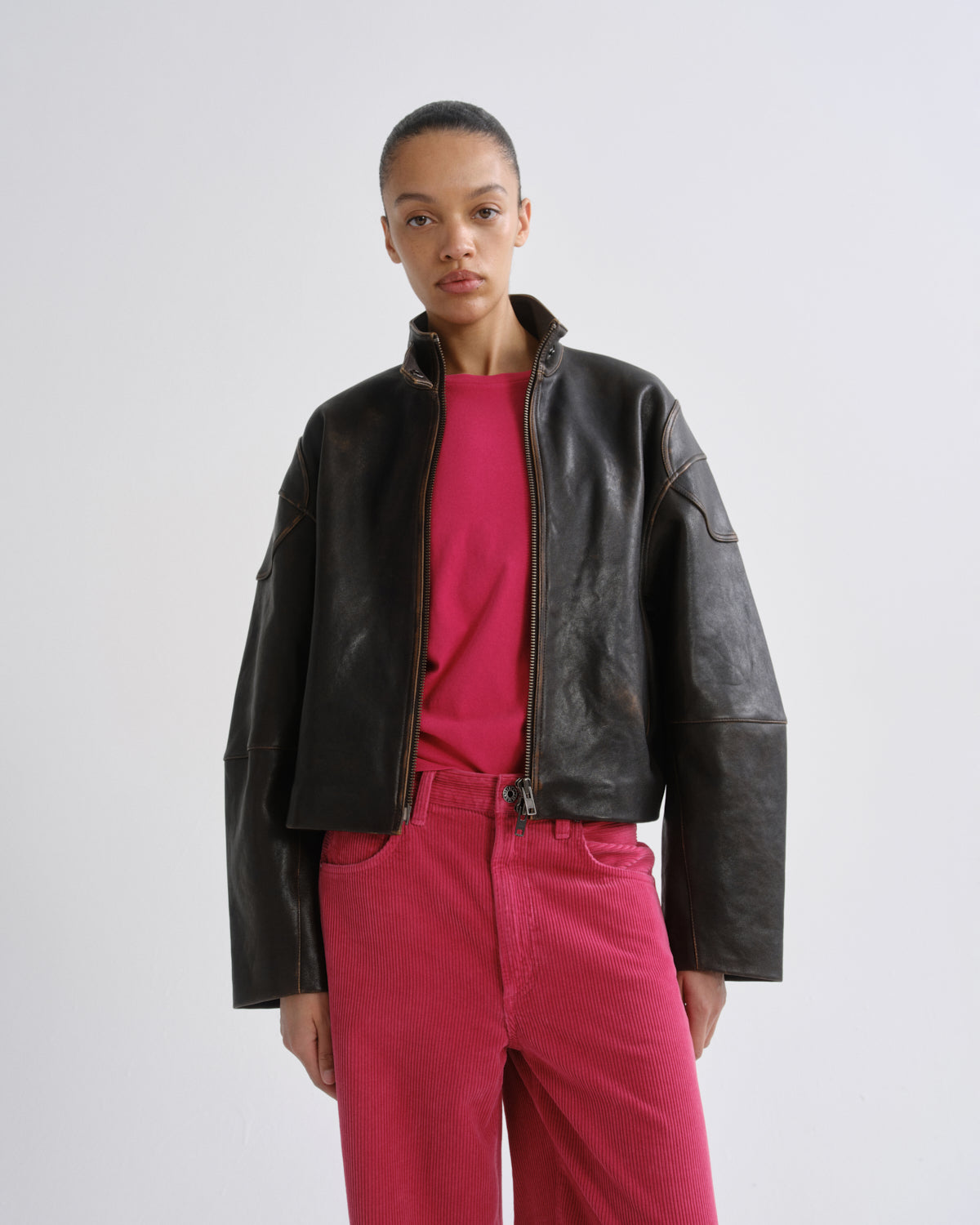 A model wearing a black leather moto jacket over a bright pink t-shirt and matching pink pants, standing against a light gray studio background.