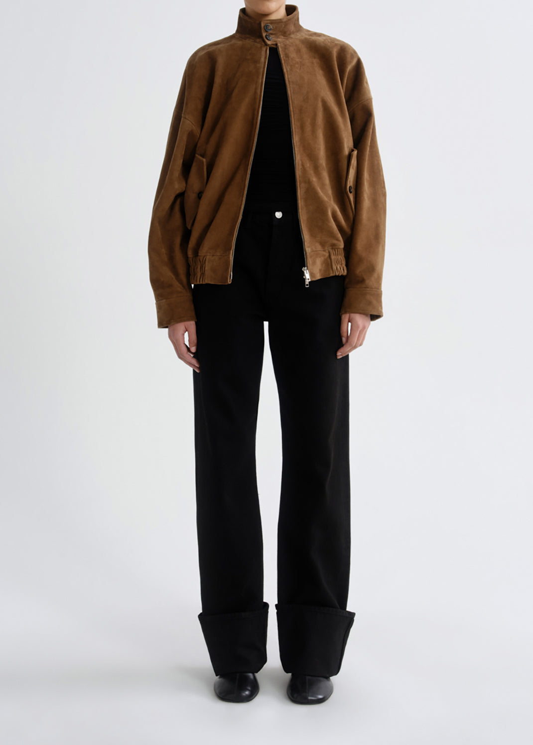 A model wearing a brown corduroy jacket with a high collar, black trousers with rolled cuffs, and black shoes, photographed against a grey background.
