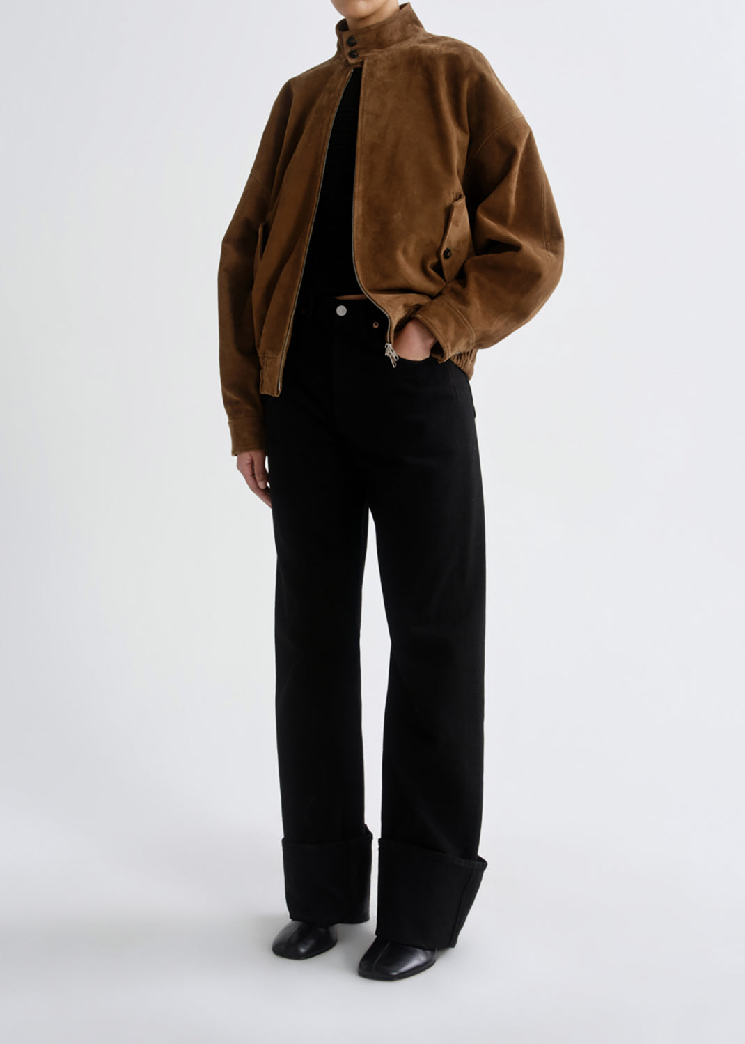 A model wearing a brown suede jacket over a black top and black wide-leg trousers, standing against a grey background.