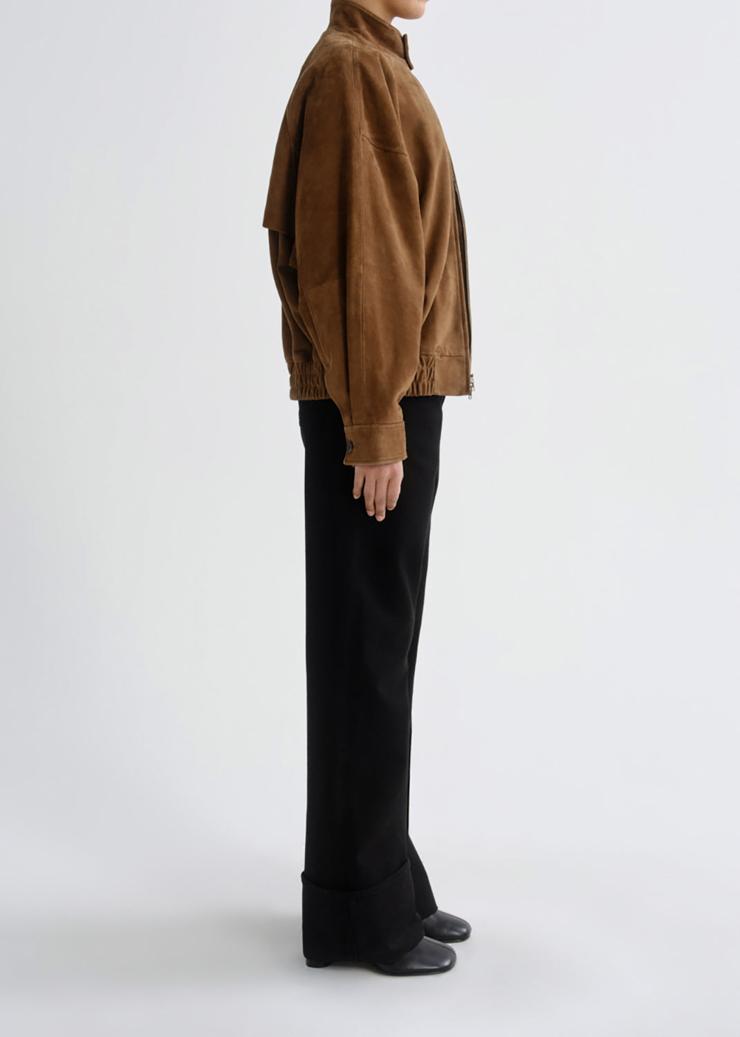 A model shown in profile wearing a brown suede jacket and black wide-leg trousers against a grey background.