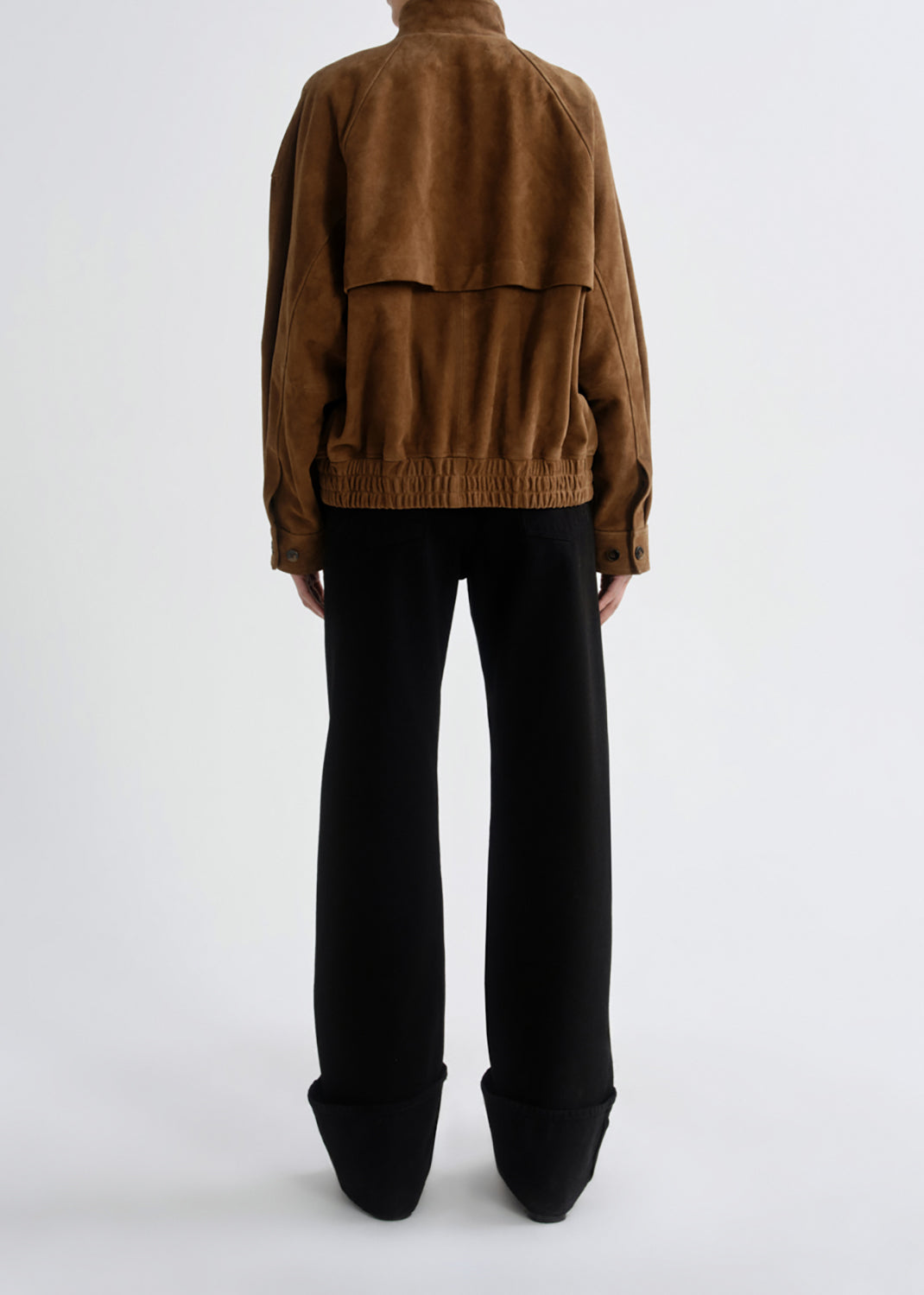A model shown from the back wearing a brown suede jacket with a draped flap detail and black wide-leg trousers, set against a grey background.