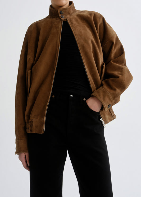 A person wearing a brown suede jacket over a black top and black pants, photographed against a grey background.