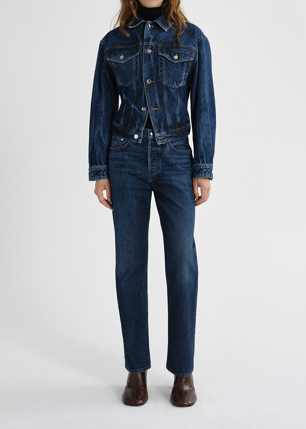 A model wearing a dark blue denim jacket and matching dark blue straight-leg jeans, paired with brown leather boots, against a grey background.