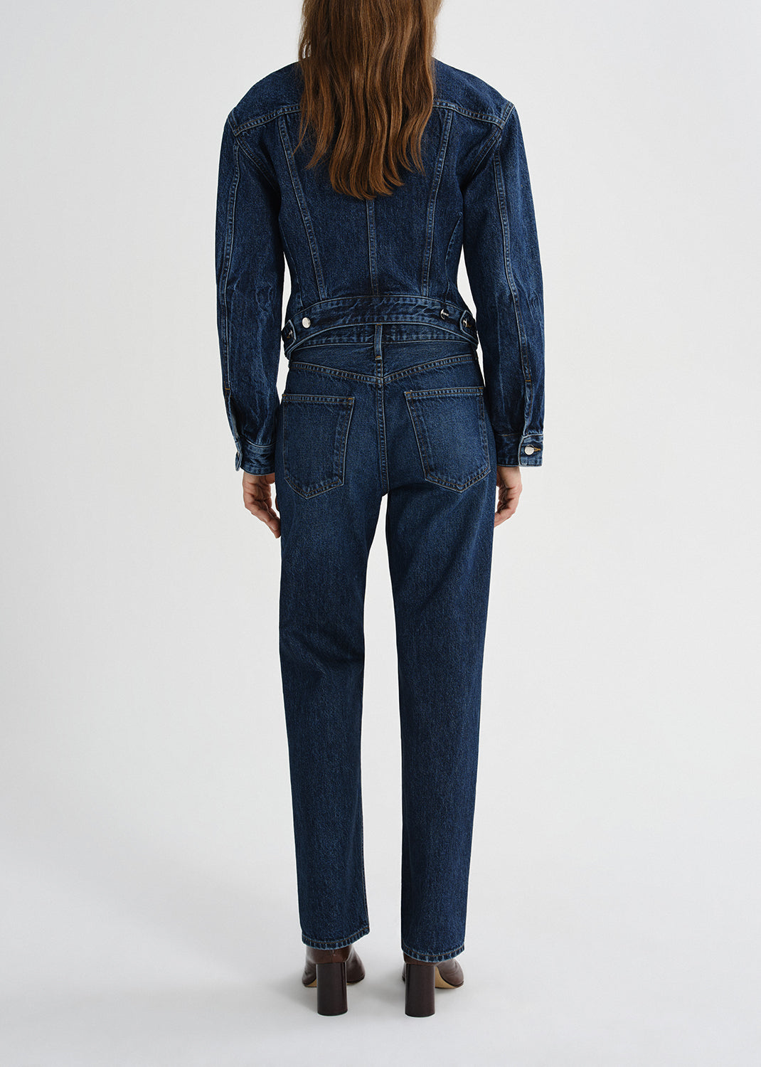 A model viewed from the back wearing a dark blue denim jumpsuit with long sleeves, a belted waist, and straight-leg pants, paired with brown block-heel shoes against a grey background.