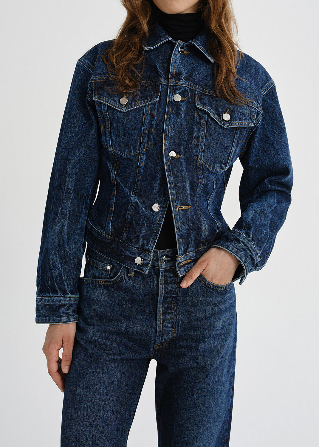 A cropped dark blue denim jacket with silver buttons, worn by a model paired with matching dark blue jeans and a black turtleneck against a grey background.