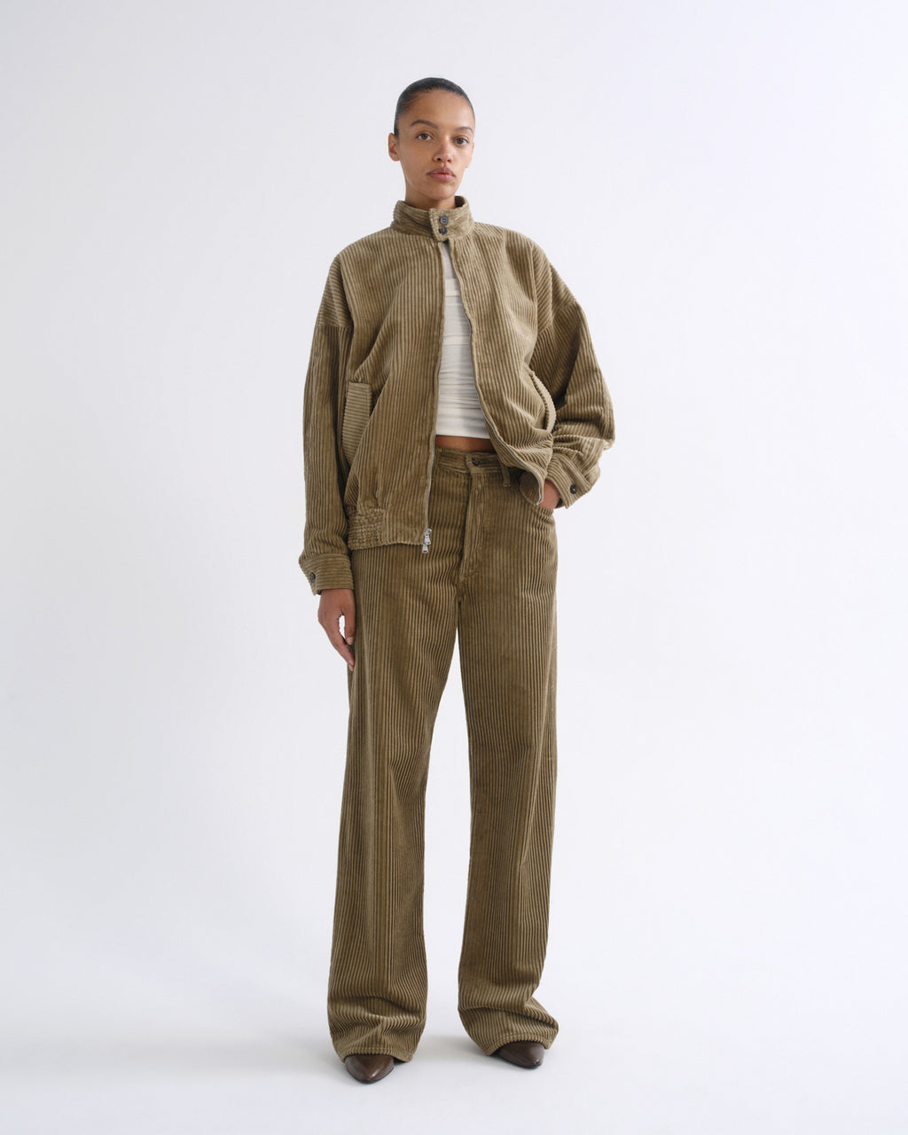 A model stands against a plain white studio background wearing a matching corduroy jacket and wide-leg pants set in a warm brownish-green color, styled with a white striped crop top.