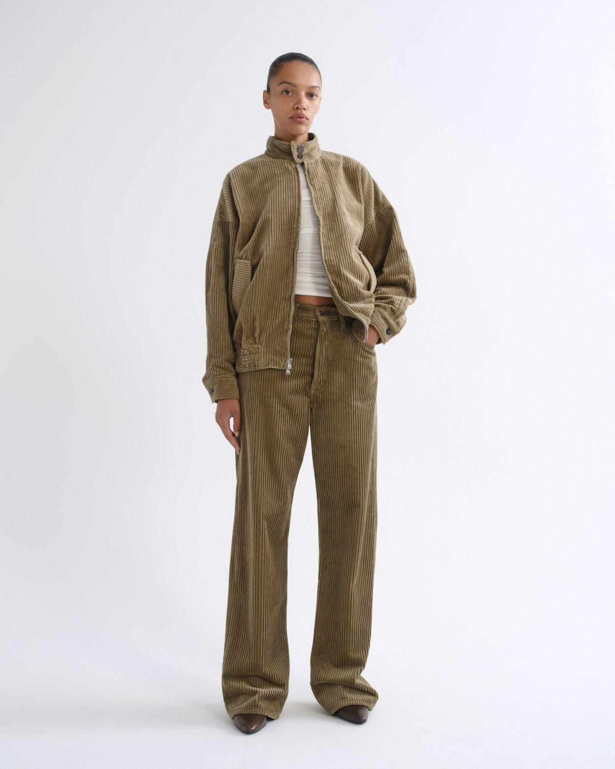 A model stands against a plain white studio background wearing a matching corduroy jacket and wide-leg pants set in a warm brownish-green color, styled with a white striped crop top.